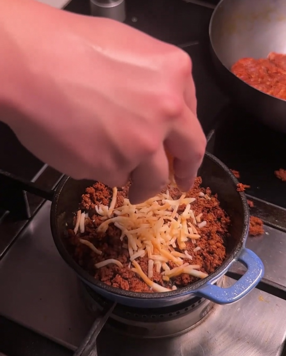 Queso rallado siendo esparcido sobre la carne de chorizo cocinada en una pequeña olla azul en la estufa.