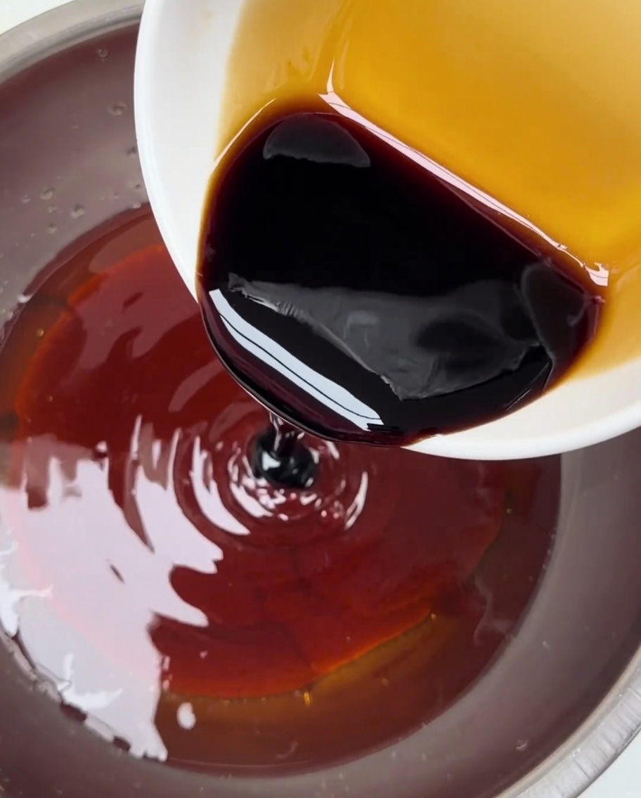 Salsa de soja oscura vertiéndose desde un pequeño cuenco en un recipiente más grande que contiene la mezcla de pasta de chile rojo.