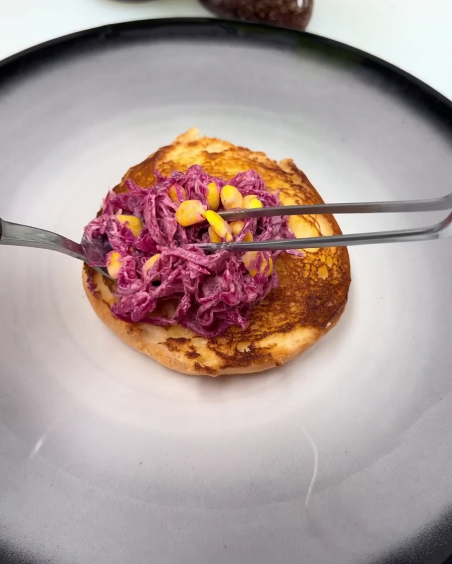 La mitad inferior de un pan de hamburguesa tostado, untada con salsa y cubierta con una generosa porción de ensalada de col lombarda y maíz.