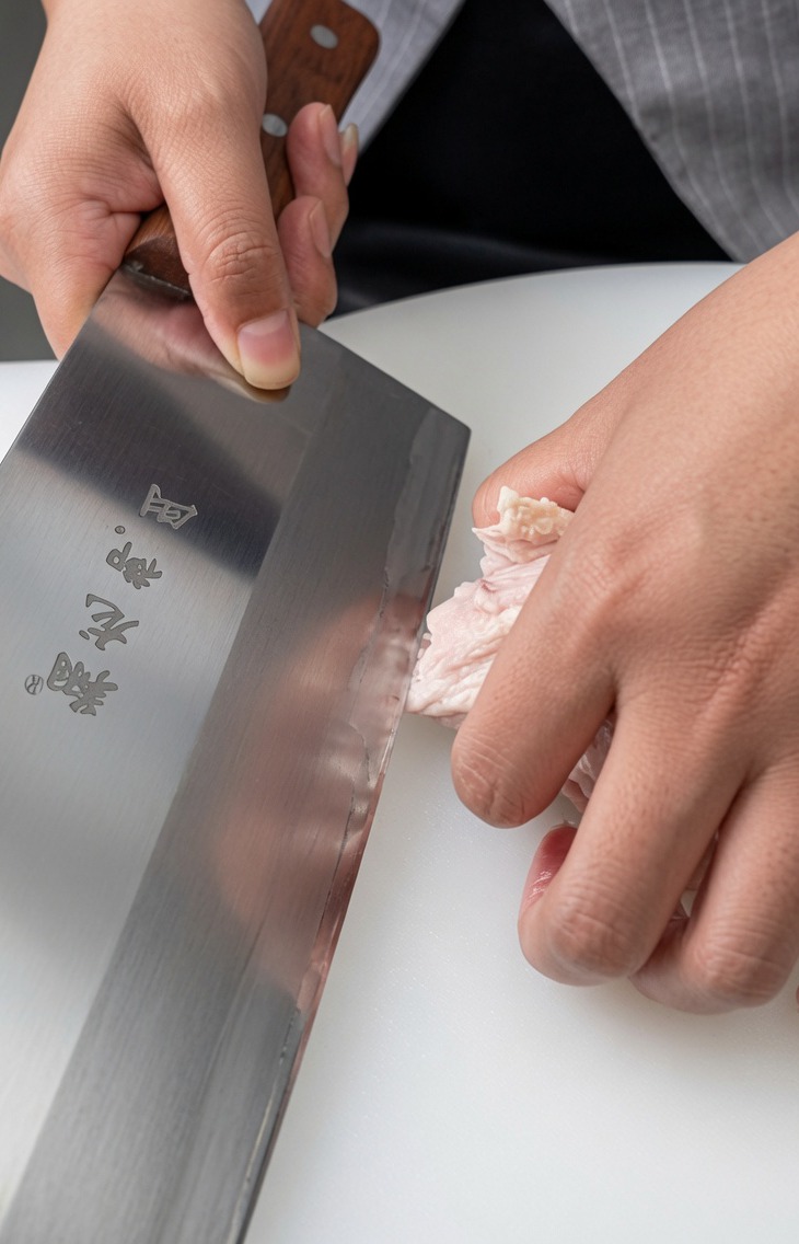Primer plano de un muslo de pollo crudo siendo deshuesado sobre una superficie blanca con un cuchillo de carnicero.