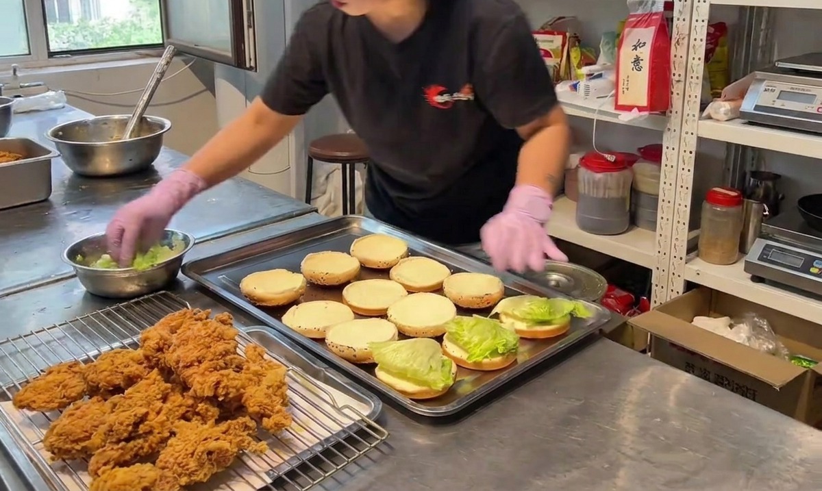 Persona colocando un trozo de pollo frito crujiente sobre la mitad inferior del pan de hamburguesa con lechuga verde.