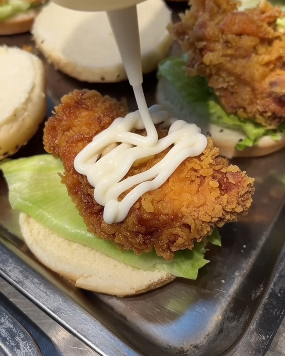 Mayonesa blanca siendo rociada desde una botella sobre un trozo de pollo frito sobre un pan de hamburguesa.