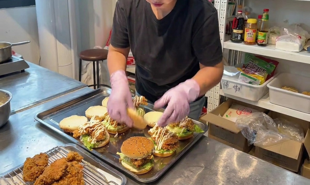 Una persona colocando la mitad superior de un pan con sésamo sobre una hamburguesa de pollo frito ya montada.