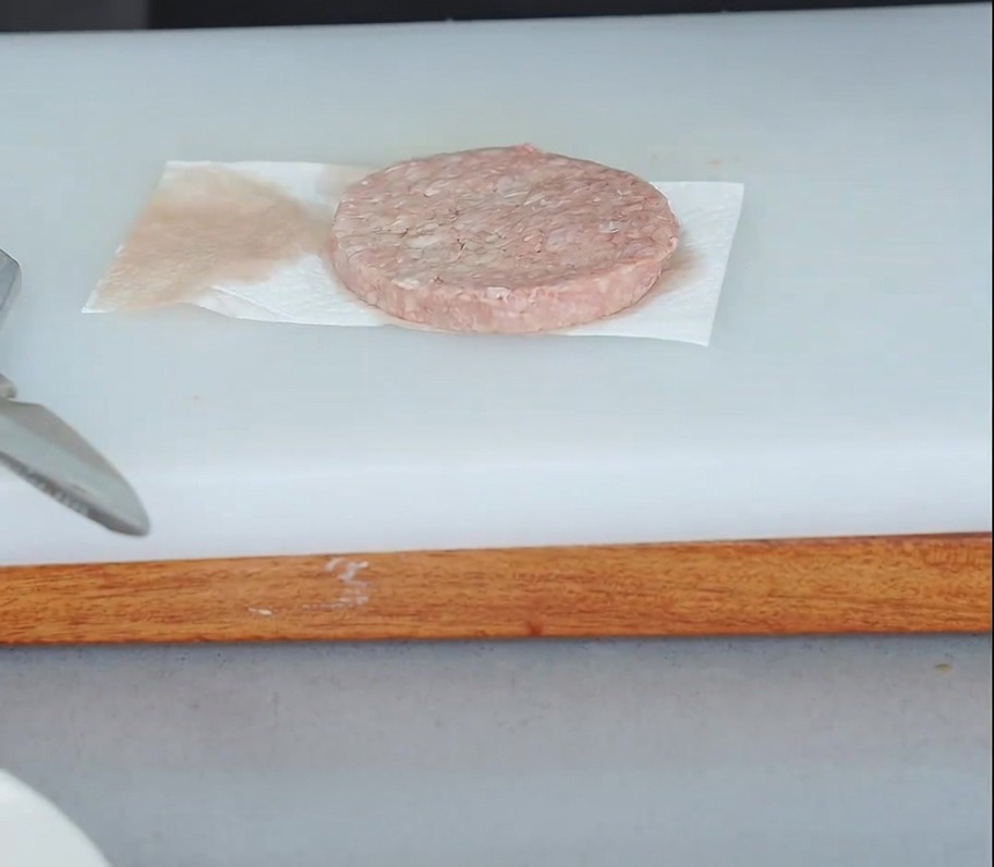 Una hamburguesa de carne cruda sobre una toalla de papel con otra toalla de papel presionada en la parte superior para absorber la humedad.