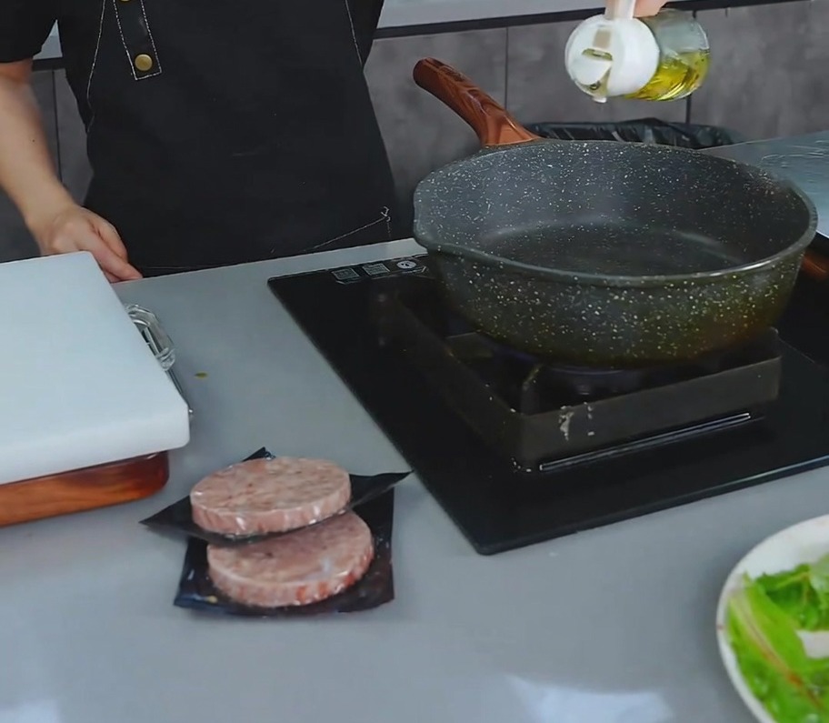 Una persona vertiendo una pequeña cantidad de aceite de cocina de una vinagrera en una sartén antiadherente oscura en la estufa.
