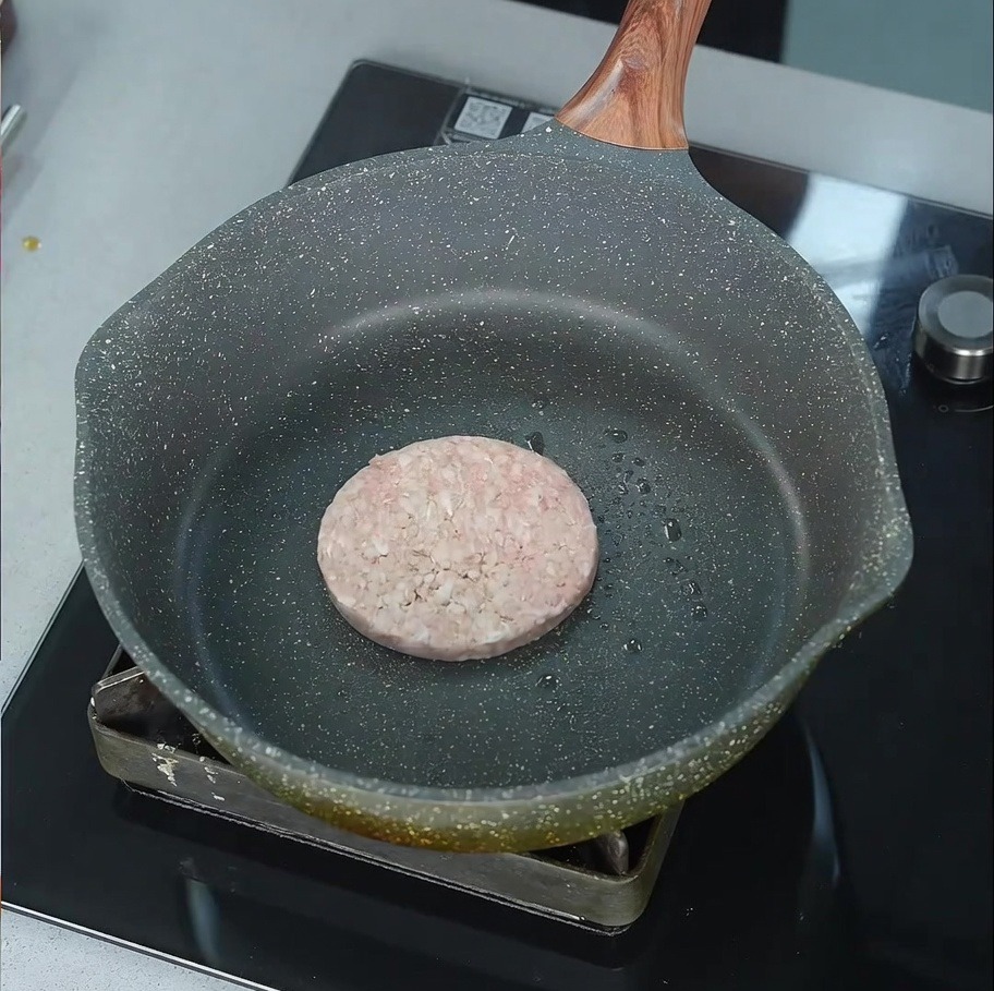 Una hamburguesa de carne cruda empezando a chisporrotear en una sartén ligeramente engrasada sobre el fuego.