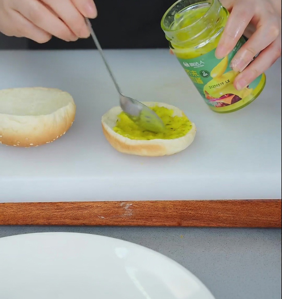 Una persona usando una cuchara para esparcir salsa relish amarilla en la mitad inferior de un pan de hamburguesa tostado.