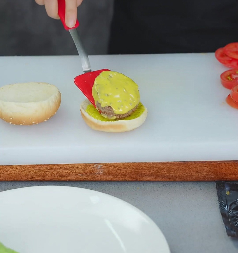 Una espátula roja levantando una hamburguesa de ternera con queso y poniéndola sobre un pan tostado cubierto de salsa relish amarilla.
