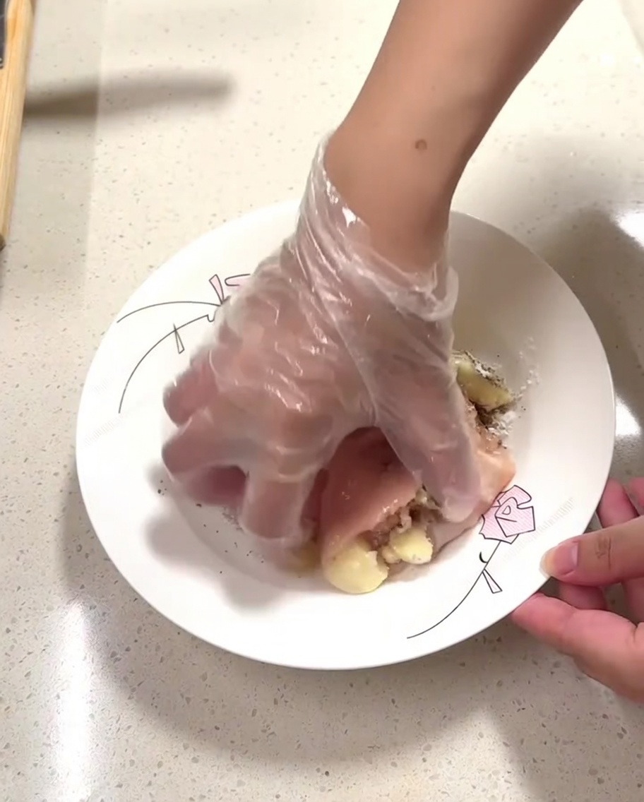 Una mano con guante masajeando los condimentos en trozos de pollo crudo en un plato.
