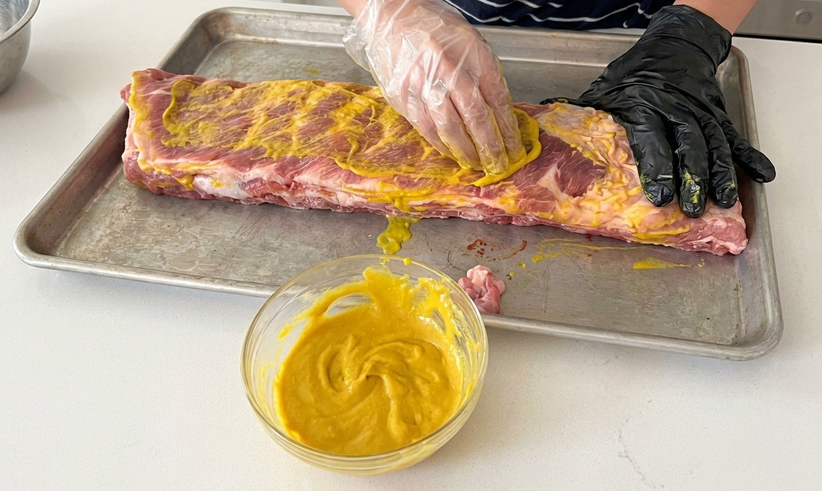 Persona usando guantes aplicando la mezcla de mostaza sobre costillas crudas en una bandeja.
