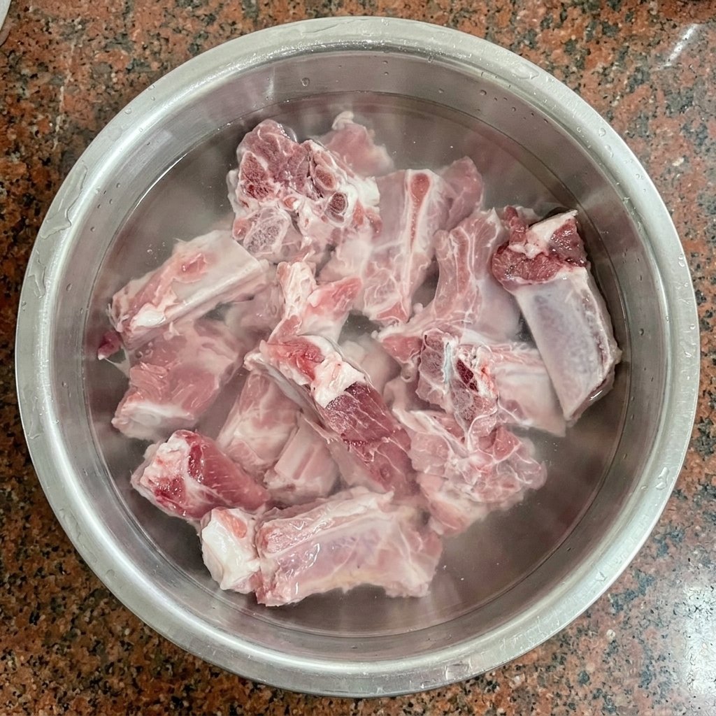 Costillas de cerdo crudas en remojo en agua fría dentro de un bol de acero inoxidable.