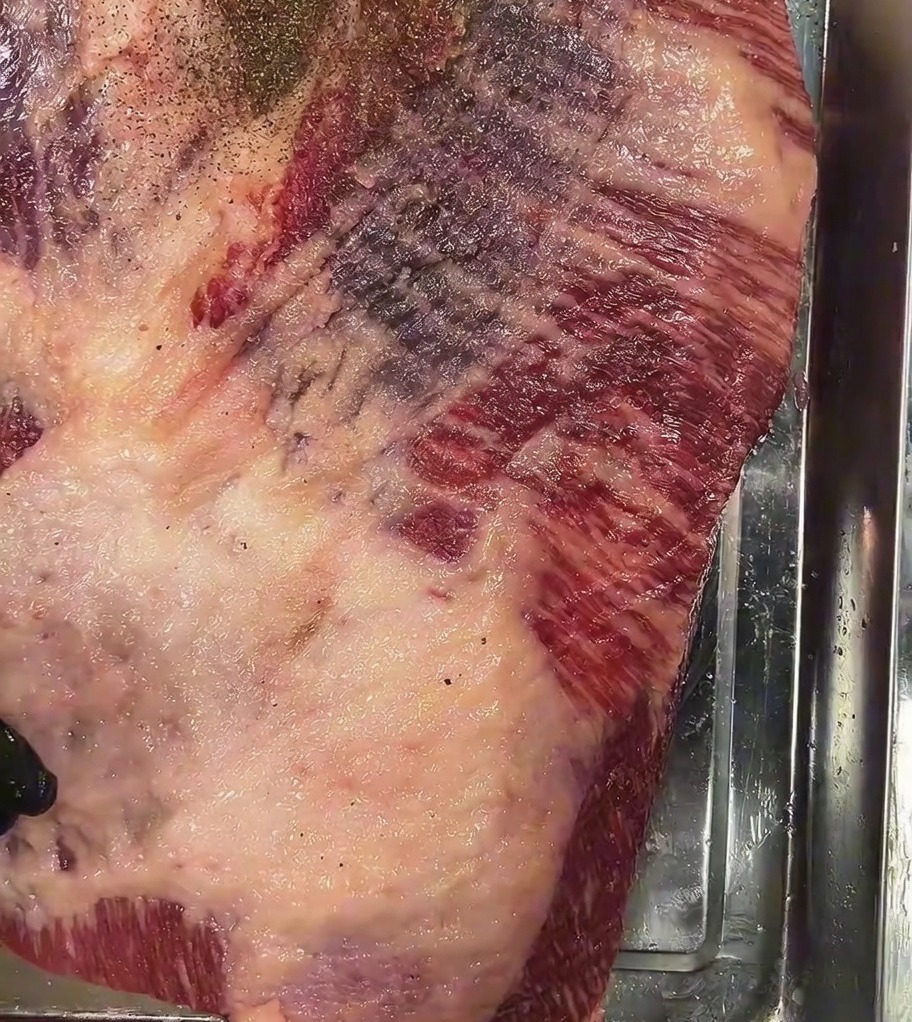 Pimienta negra gruesa espolvoreada generosamente sobre la superficie salada del brisket crudo.
