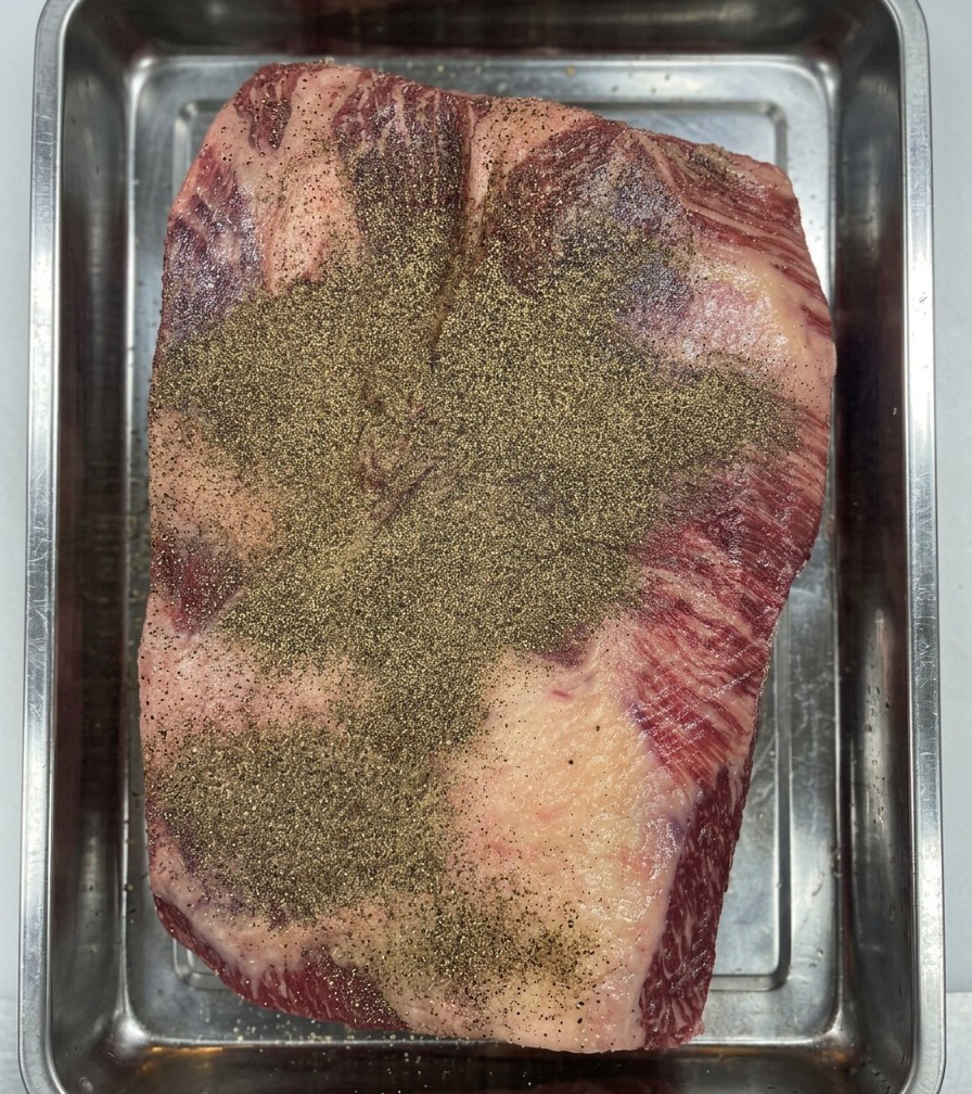 Manos enguantadas presionando la pimienta negra en la superficie del brisket Wagyu para crear una capa uniforme.