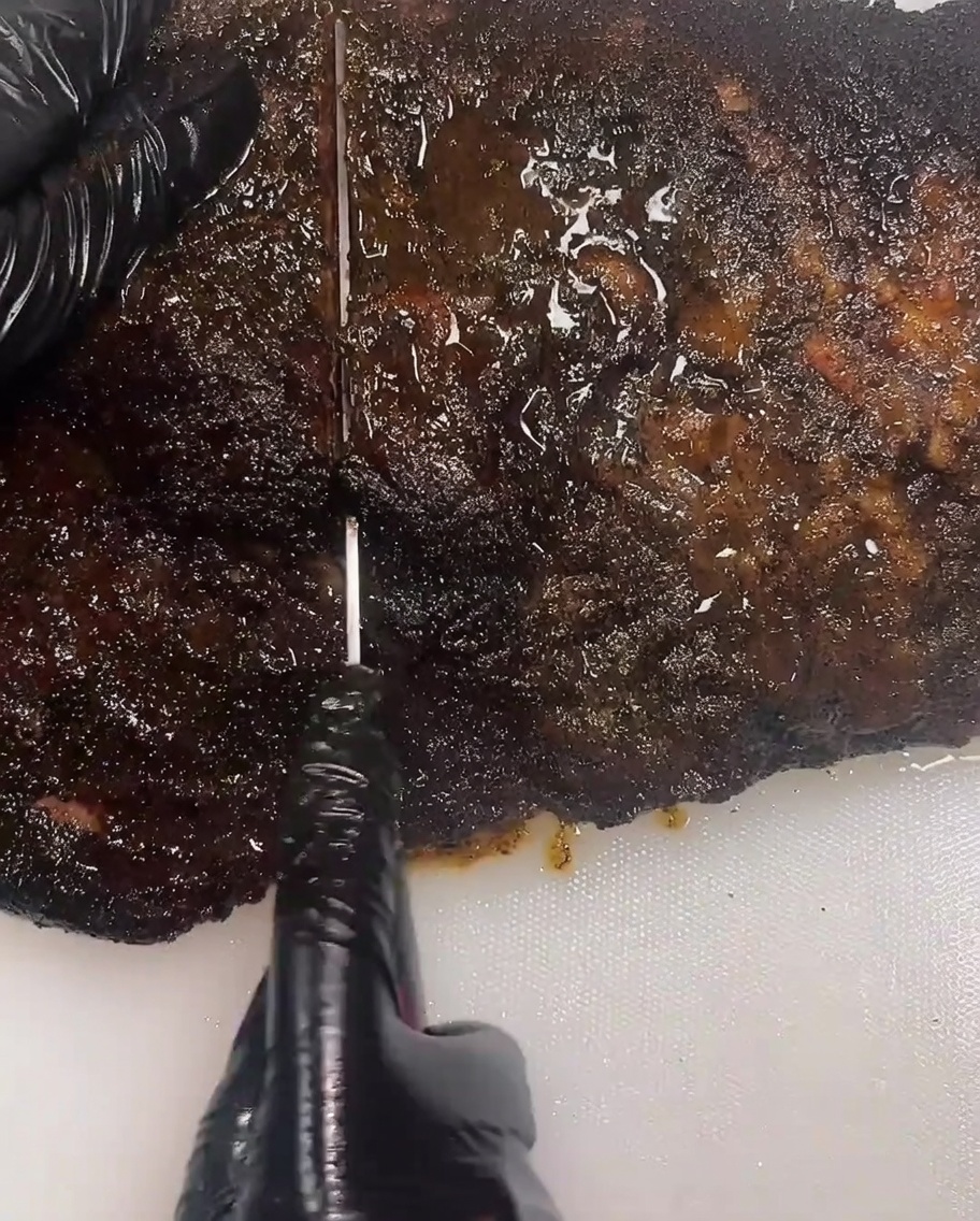 Primer plano de una persona usando guantes negros con un cuchillo largo para rebanar un brisket ahumado oscuro sobre una tabla de cortar blanca.