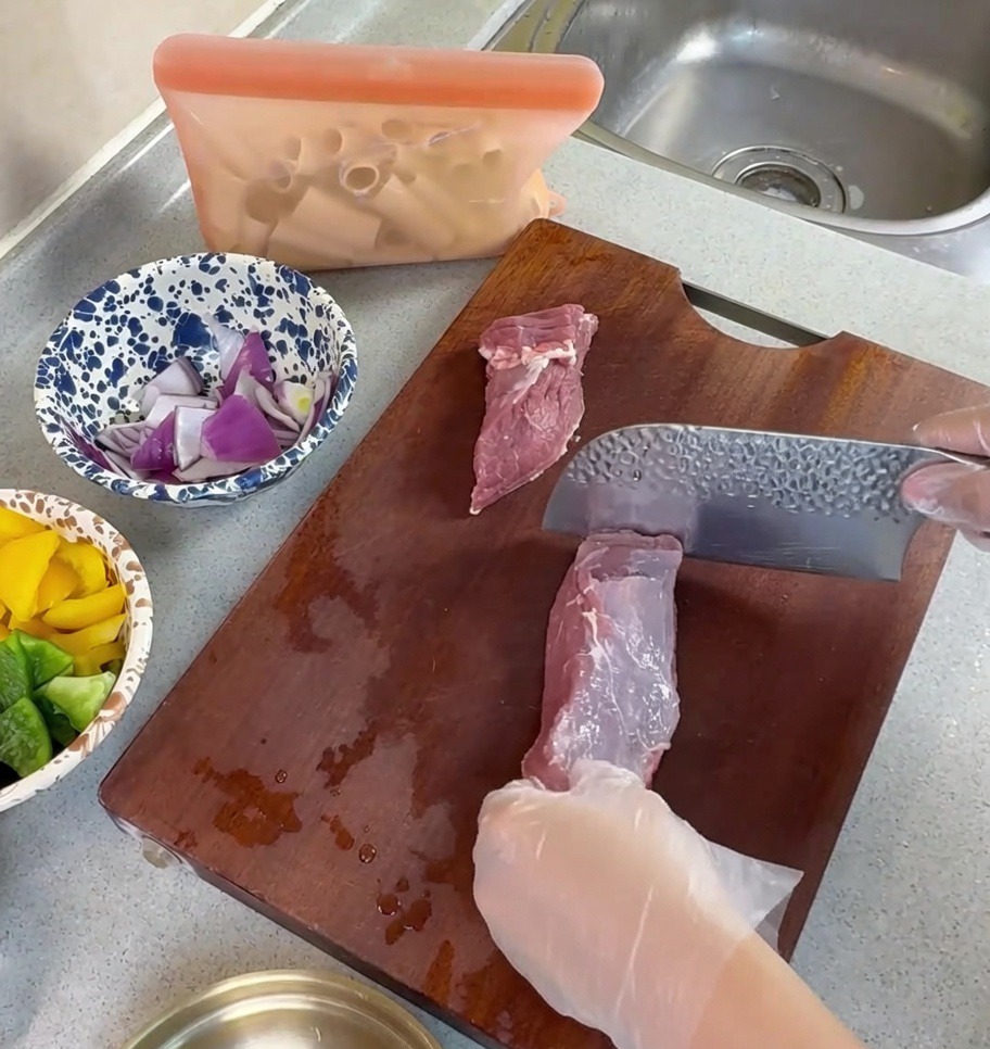 Manos con guantes usando un cuchillo para cortar cerdo crudo en tiras finas sobre una tabla de madera.