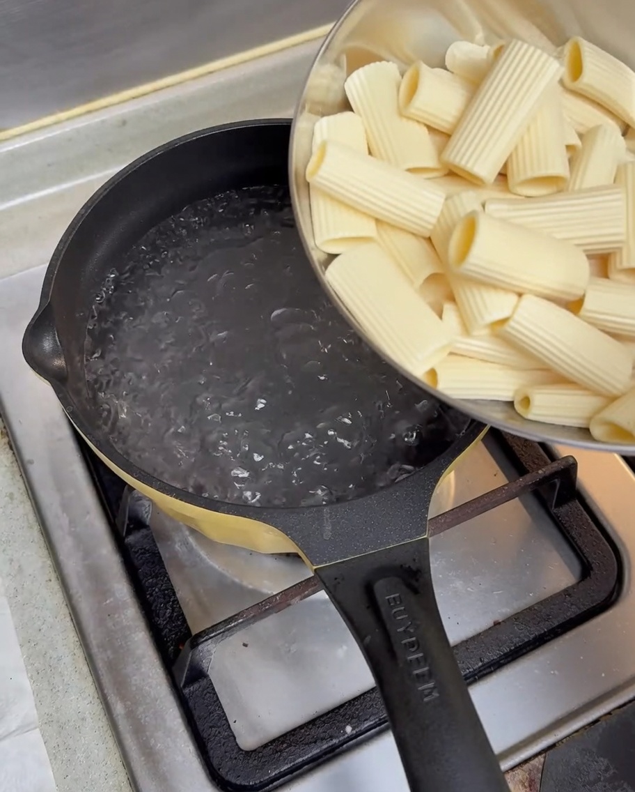 Pasta rigatoni pre-hidratada siendo vertida desde un bol en una olla con agua hirviendo.