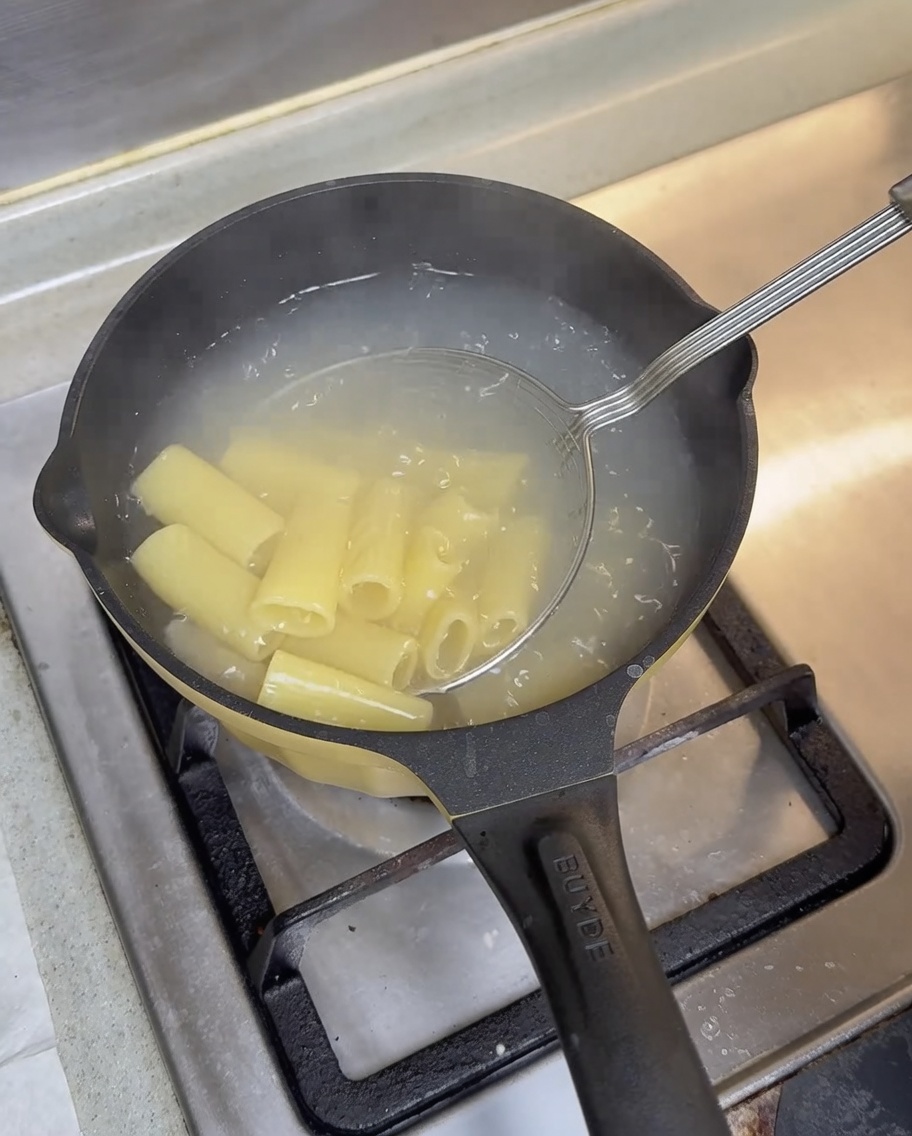 Un colador metálico retirando rigatoni cocidos del agua hirviendo.