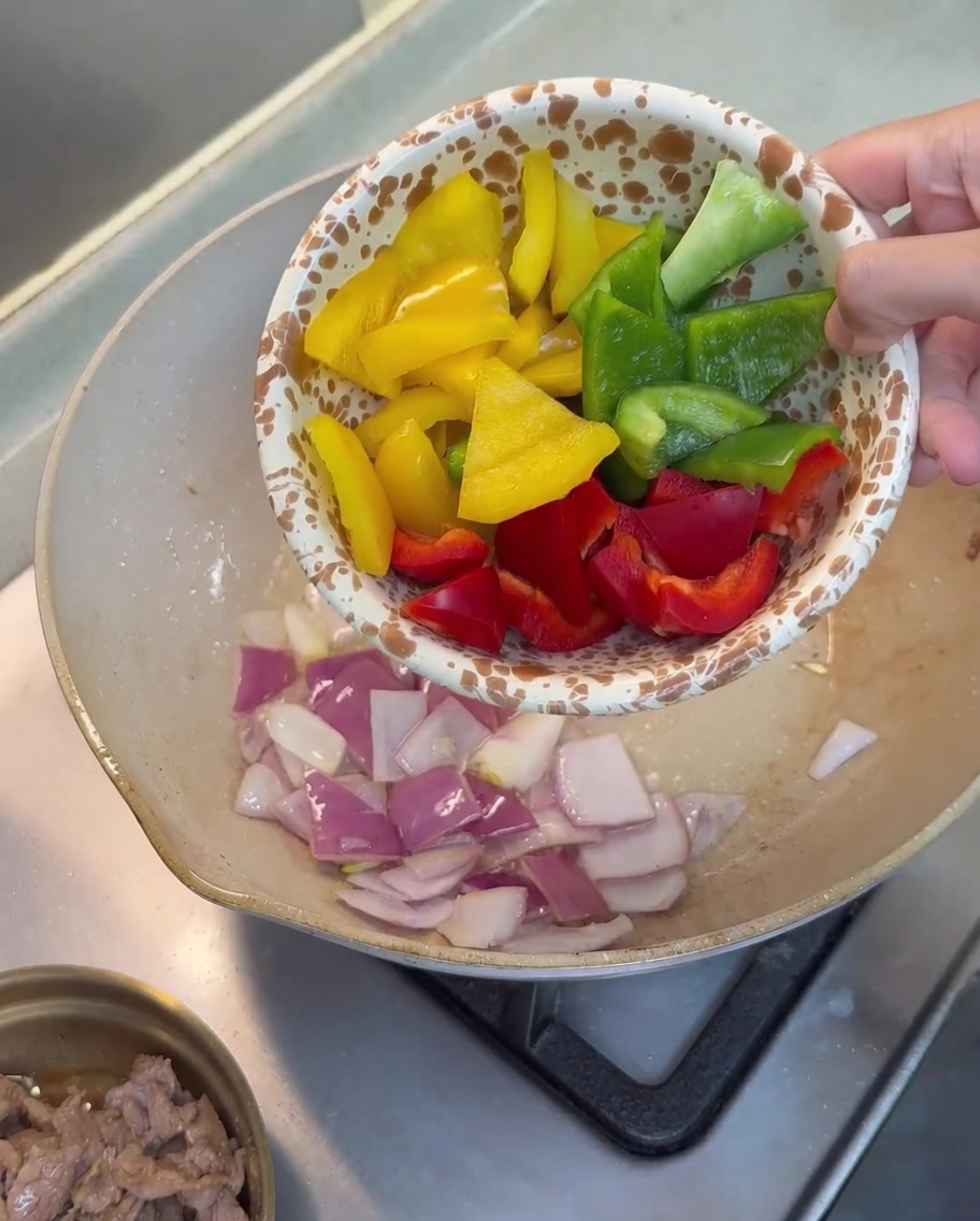Trozos de pimiento rojo, amarillo y verde siendo añadidos a la sartén con cebollas.