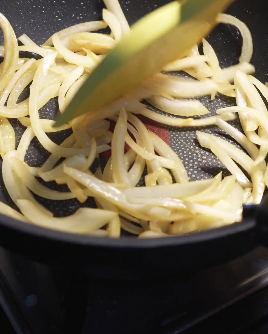 Rodajas de cebolla blanca siendo salteadas con una espátula en una sartén caliente hasta ablandarse.