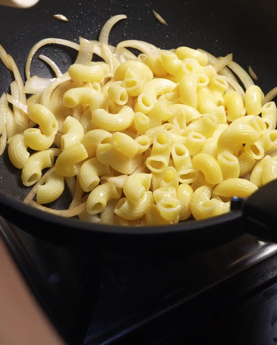 Macarrones cocidos siendo vertidos en una sartén con cebollas cortadas ablandadas.