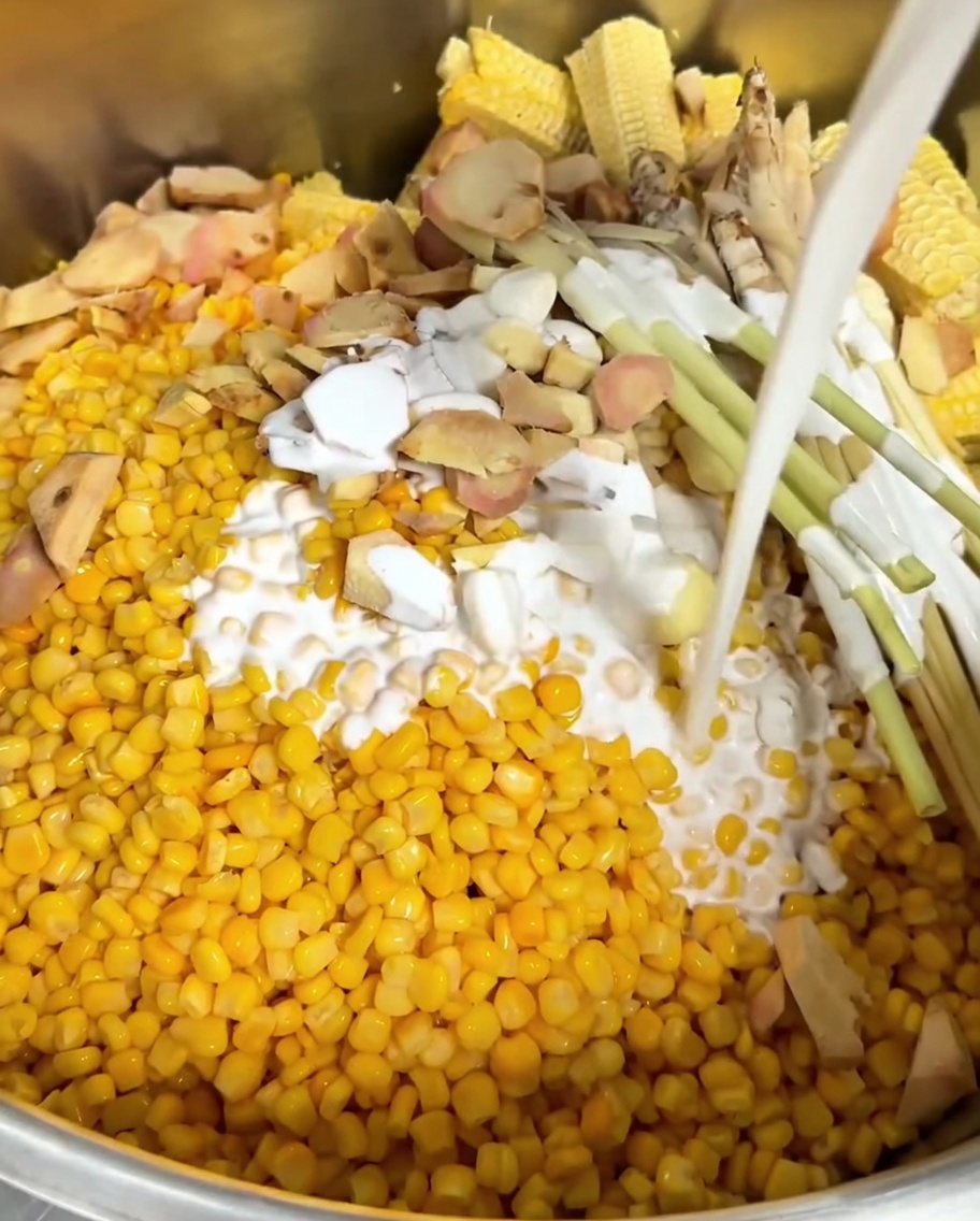Leche de coco siendo vertida sobre el elote, aromáticos y mazorcas desgranadas en la olla.