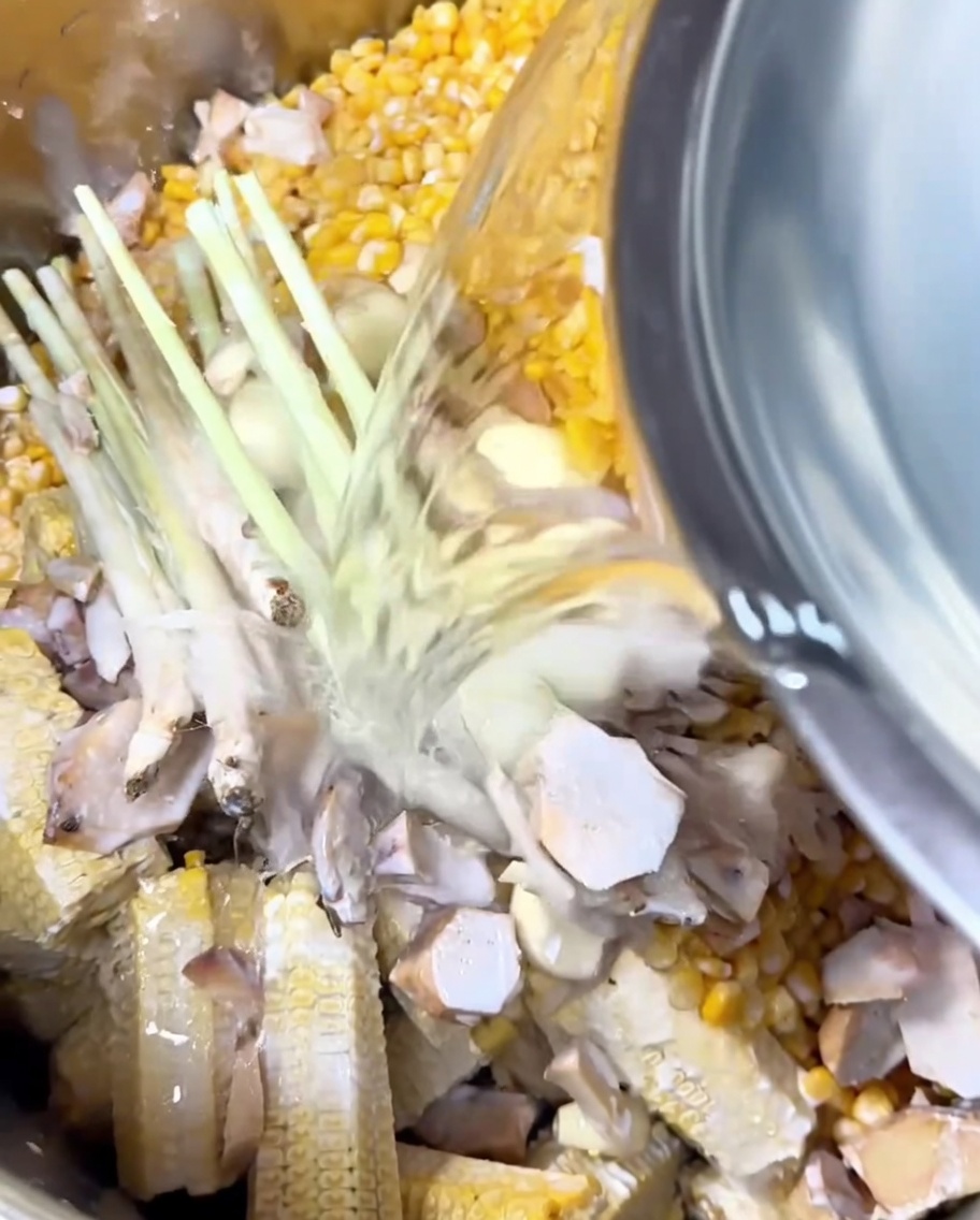 Agua clara siendo vertida de un tazón a la olla con el elote, aromáticos y leche de coco.