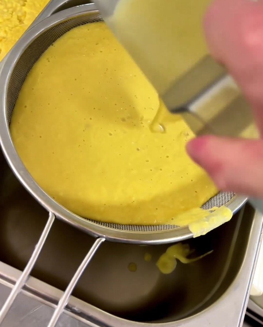 Sopa de elote amarilla siendo colada a través de un tamiz fino hacia un recipiente.