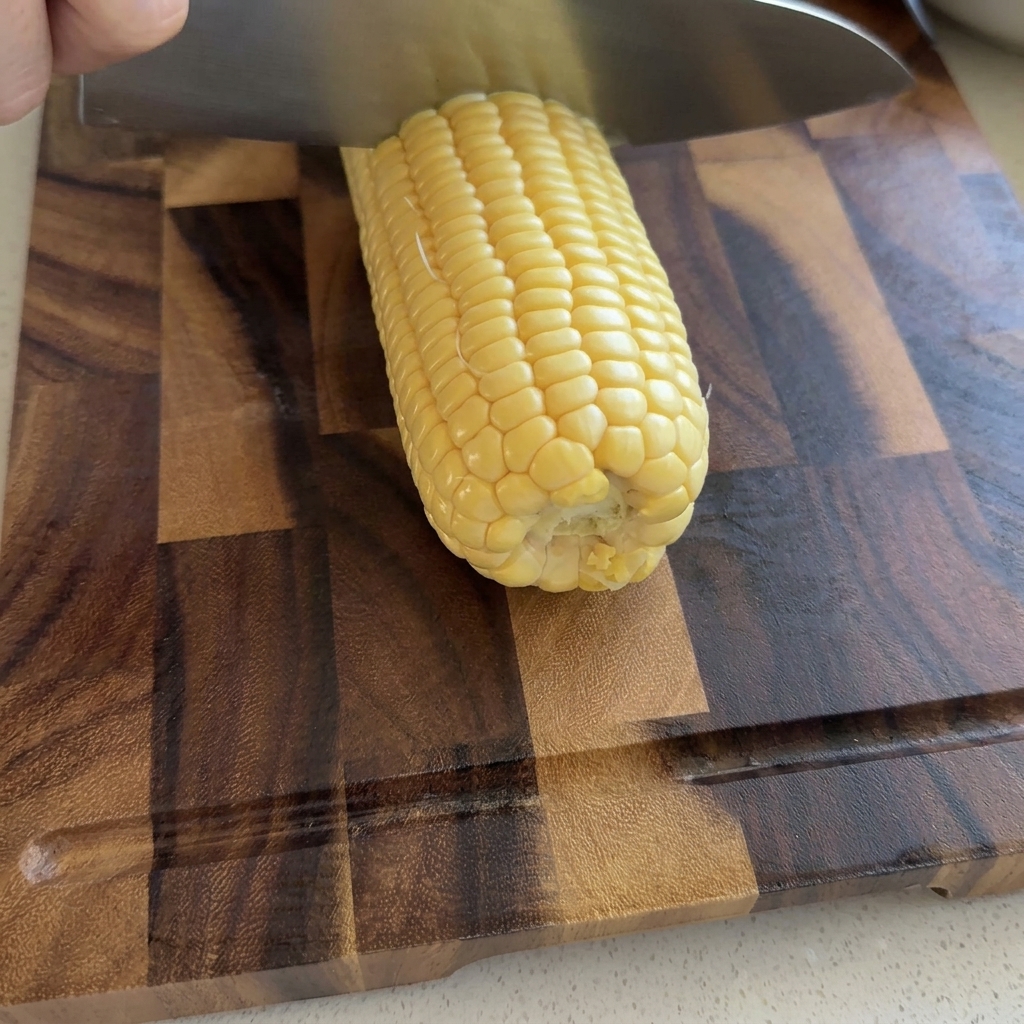 Una mano usando un cuchillo de chef grande para rebanar granos de maíz amarillo crudo de una mazorca sobre una tabla de cortar de madera.
