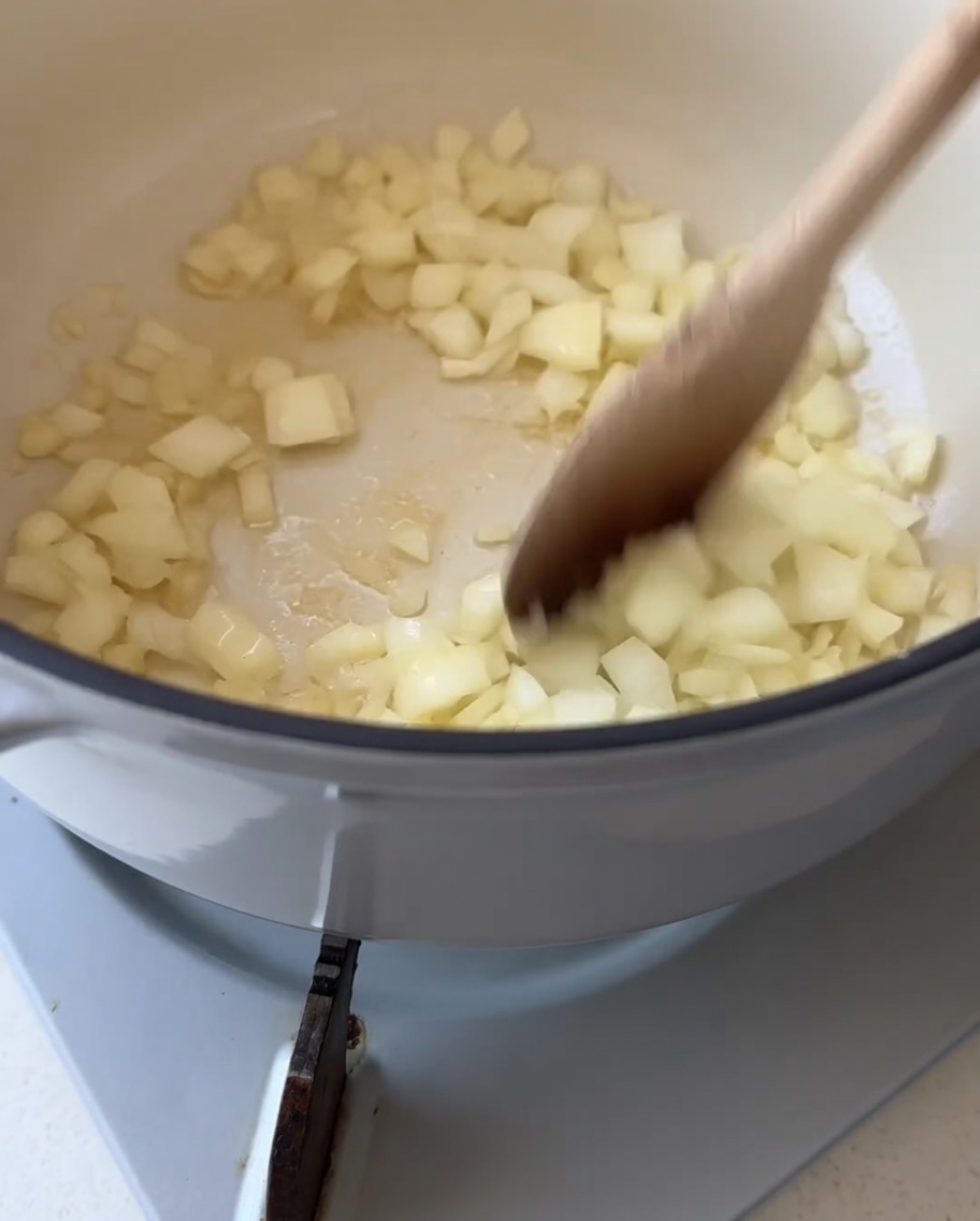 Una cuchara de madera revolviendo cebolla blanca picada dentro de una olla blanca con mantequilla derretida.