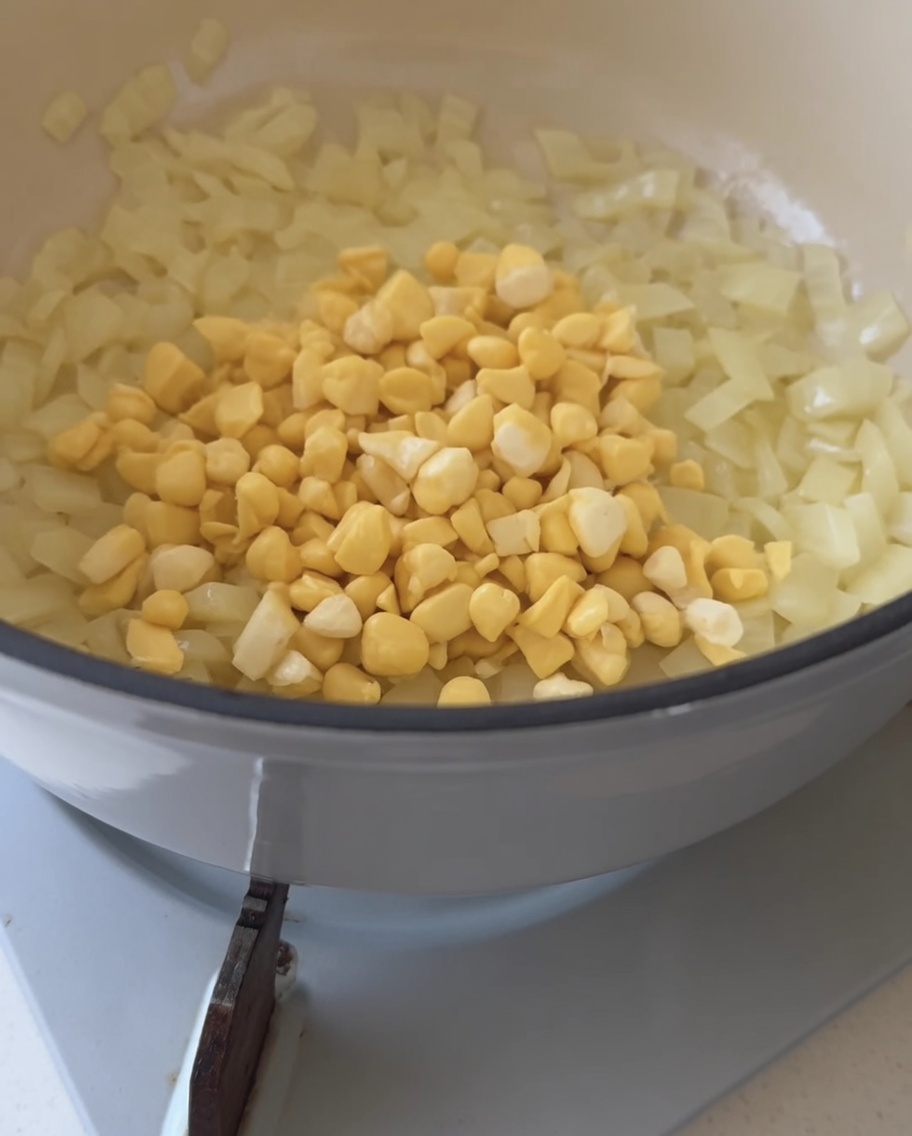 Granos frescos de maíz amarillo siendo vertidos en una olla que contiene cebollas sofritas translúcidas.