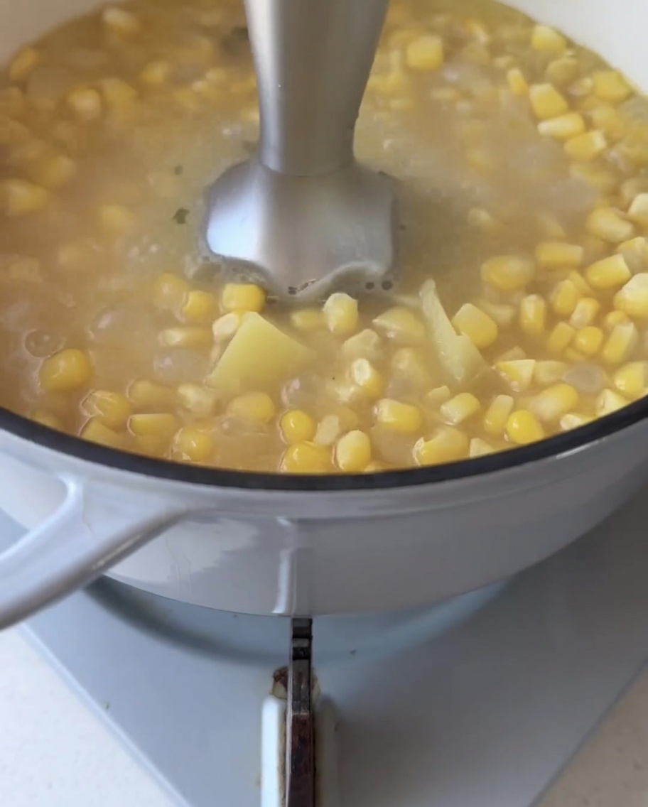 Una batidora de mano sumergida en una olla de granos de maíz y trozos de batata hirviendo a fuego lento, mezclándolos hasta obtener un líquido suave.