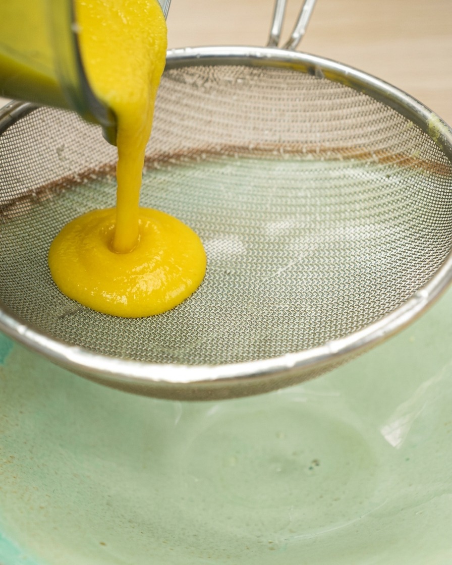 Puré espeso de maíz amarillo siendo colado a través de un colador de acero inoxidable de malla fina sobre un tazón.