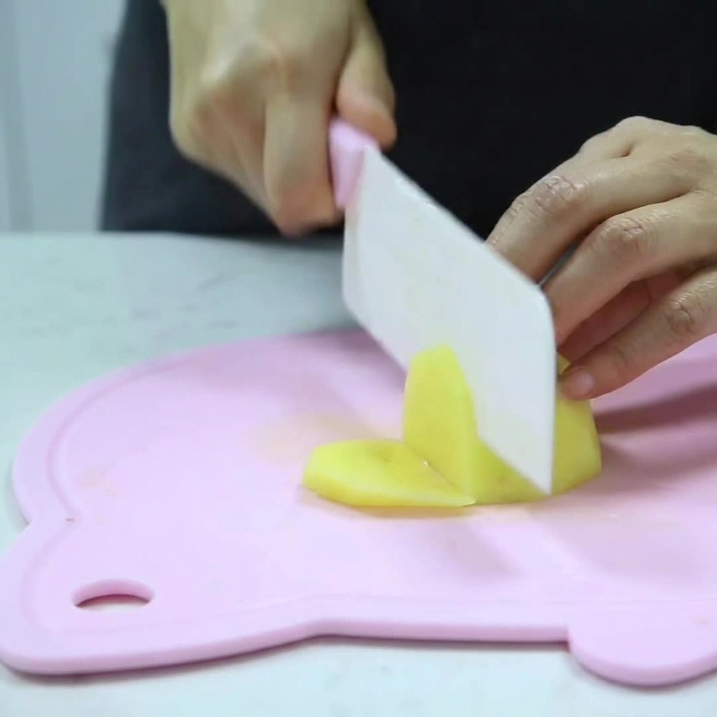 Manos sosteniendo un cuchillo, cortando una patata pelada en pequeños dados sobre una tabla de cortar rosa.