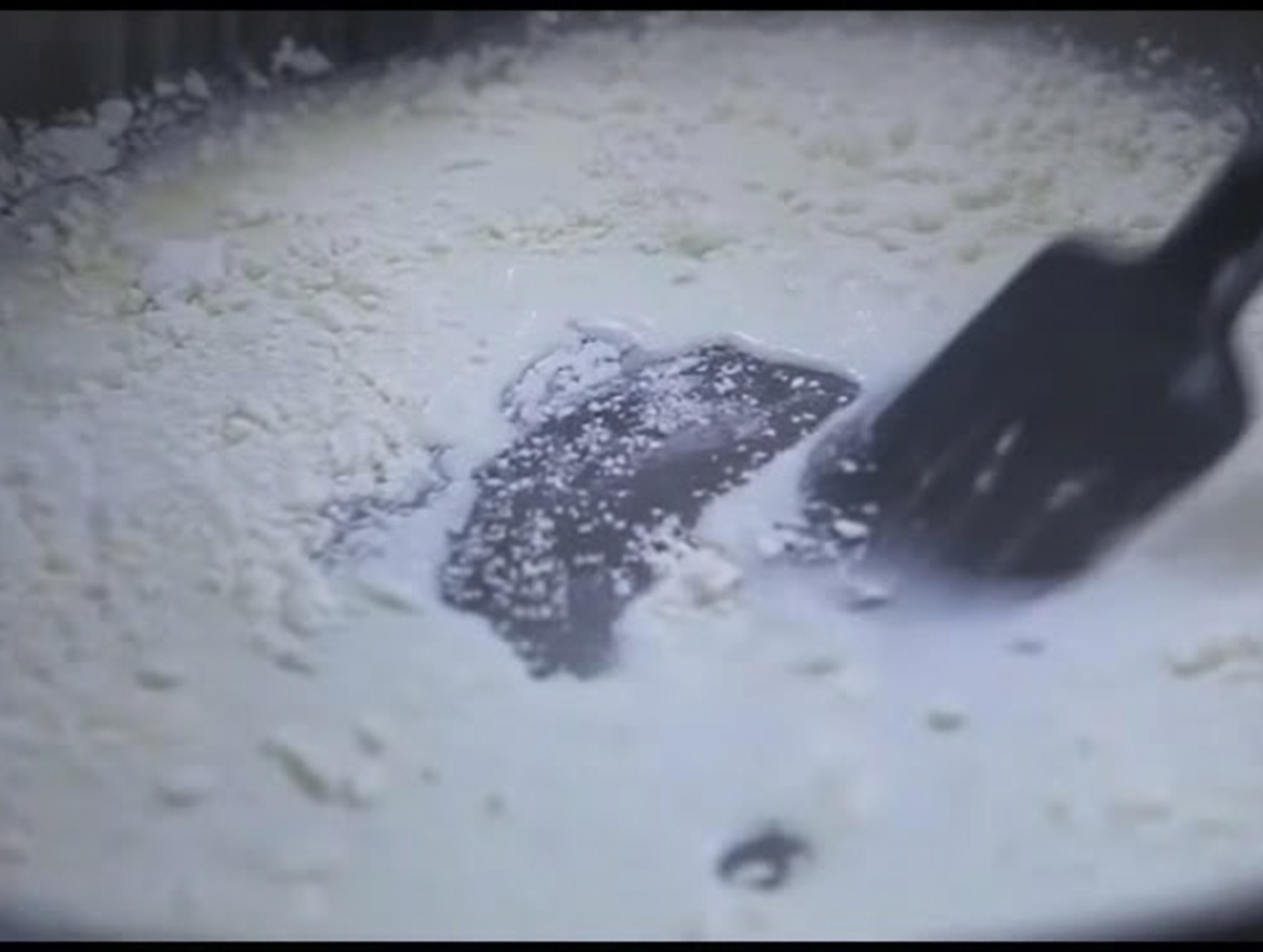 Una espátula de silicona negra removiendo una mezcla espesa y blanca de leche y harina en una sartén.