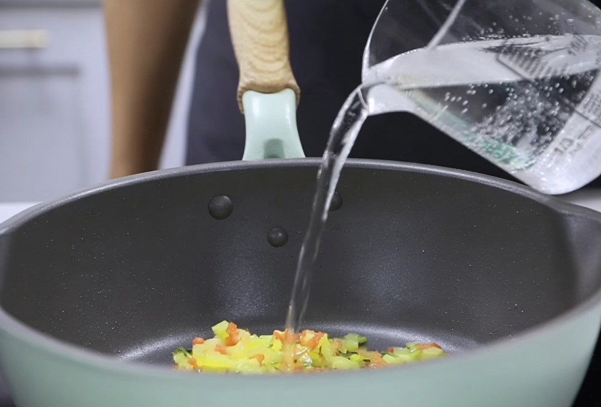 Agua vertiéndose desde una taza medidora en una sartén con verduras salteadas, preparándose para incorporar la mezcla de leche espesada.