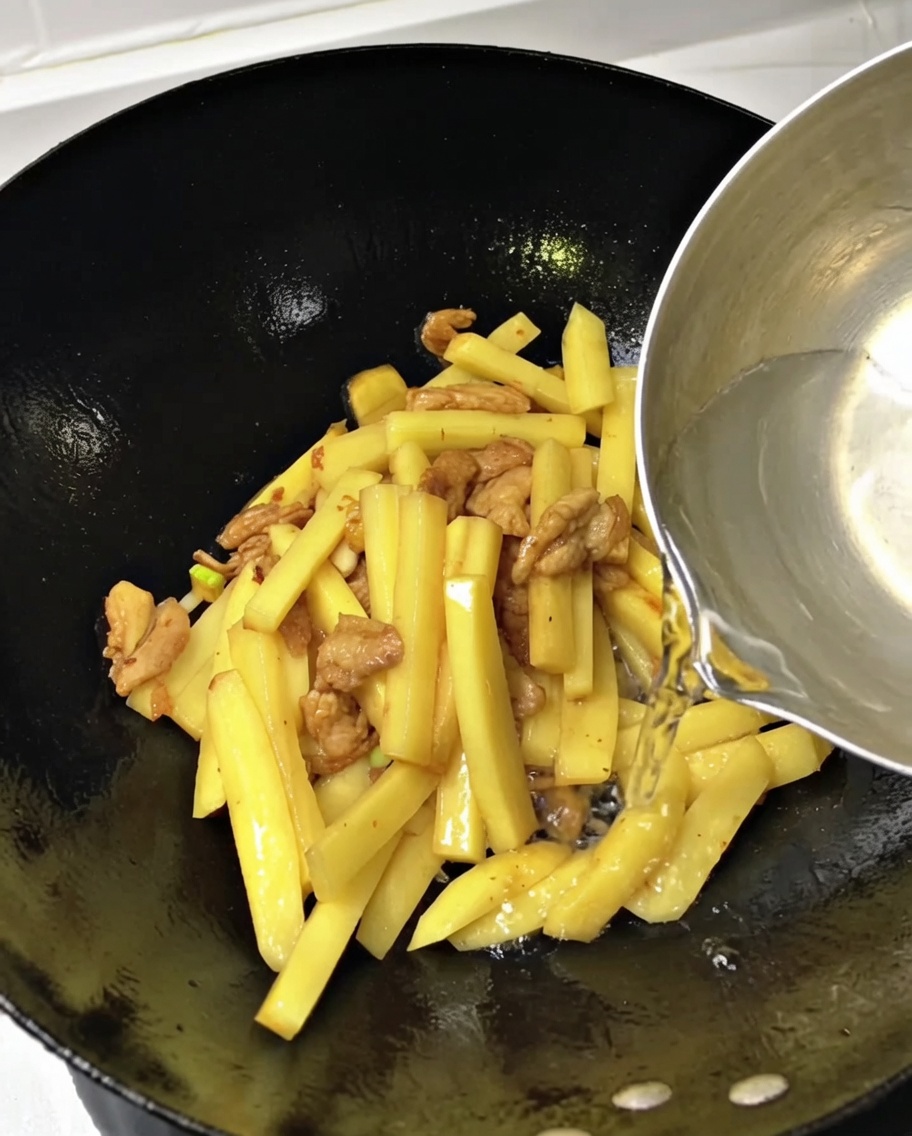 Agua siendo vertida con un cazo en un wok lleno de tiras de patata y cerdo.