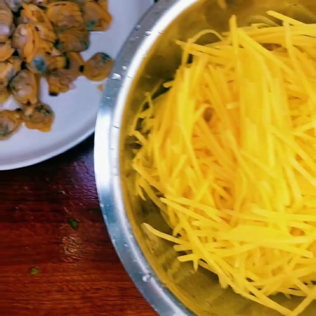 Un bol que contiene patatas crudas ralladas, carne de almeja cocida y un pequeño montón de cebolletas picadas.