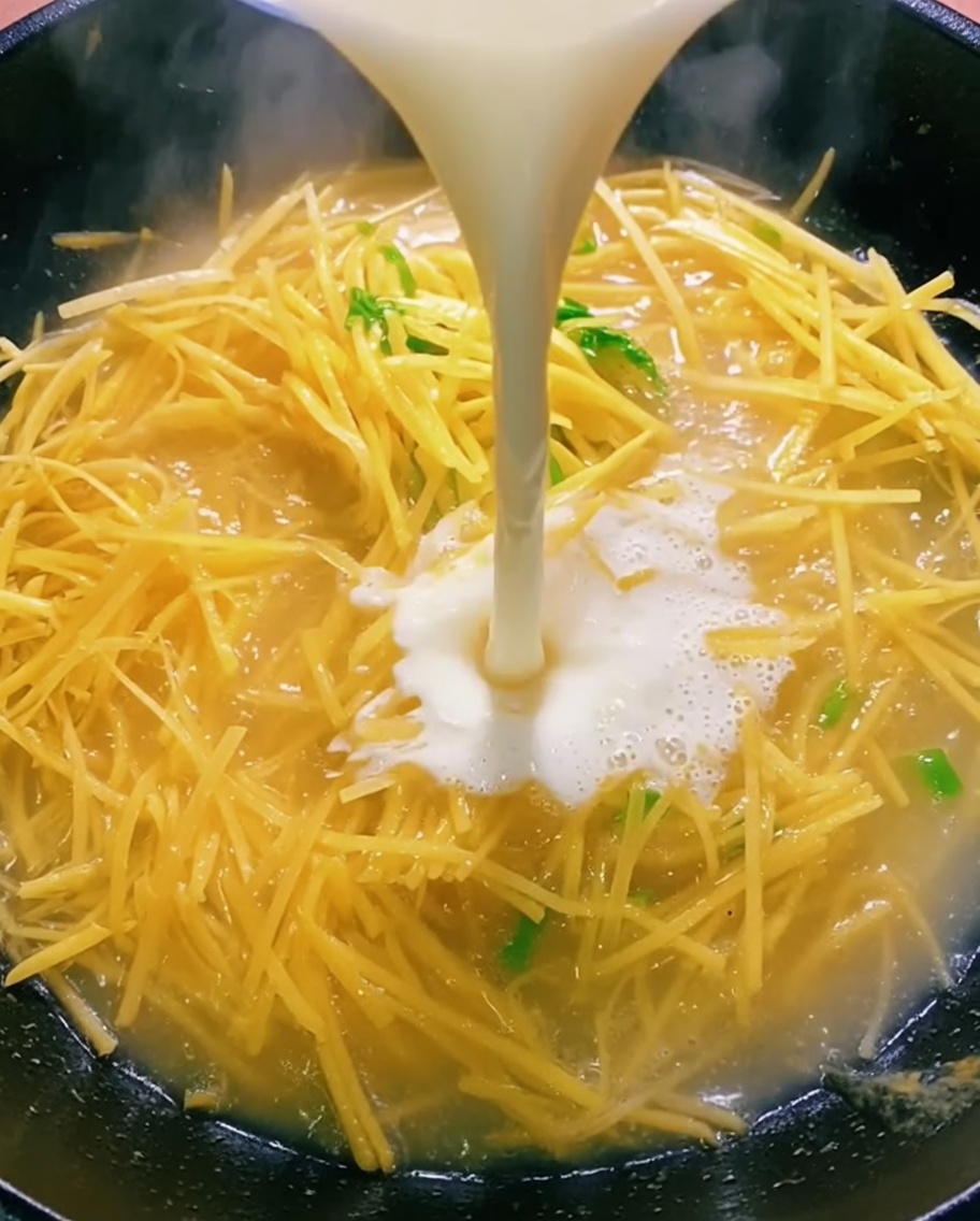 Líquido claro vertiéndose desde un bol a un wok que contiene patatas salteadas.