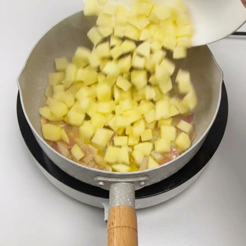 Cubos de patata deslizándose de un plato blanco a una sartén con cebolla salteada.