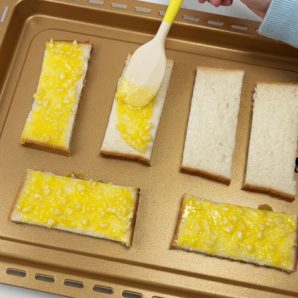Una espátula extendiendo mantequilla de ajo sobre rebanadas de pan en una bandeja de horno.