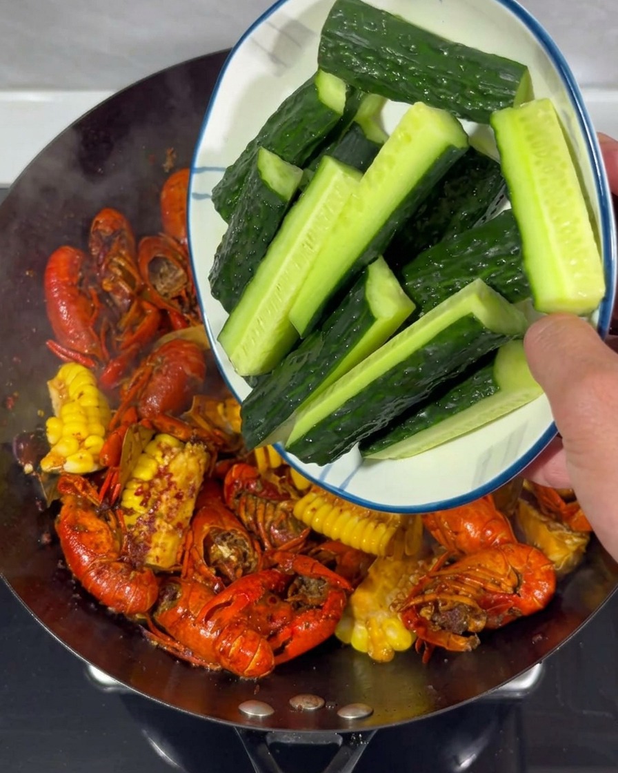 Bastones de pepino fresco siendo añadidos desde un plato blanco a un wok con cangrejos y maíz.