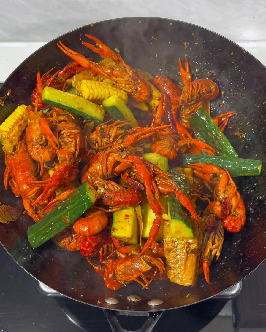 Primer plano de cangrejos, maíz y pepino siendo mezclados en un wok con una salsa roja oscura.