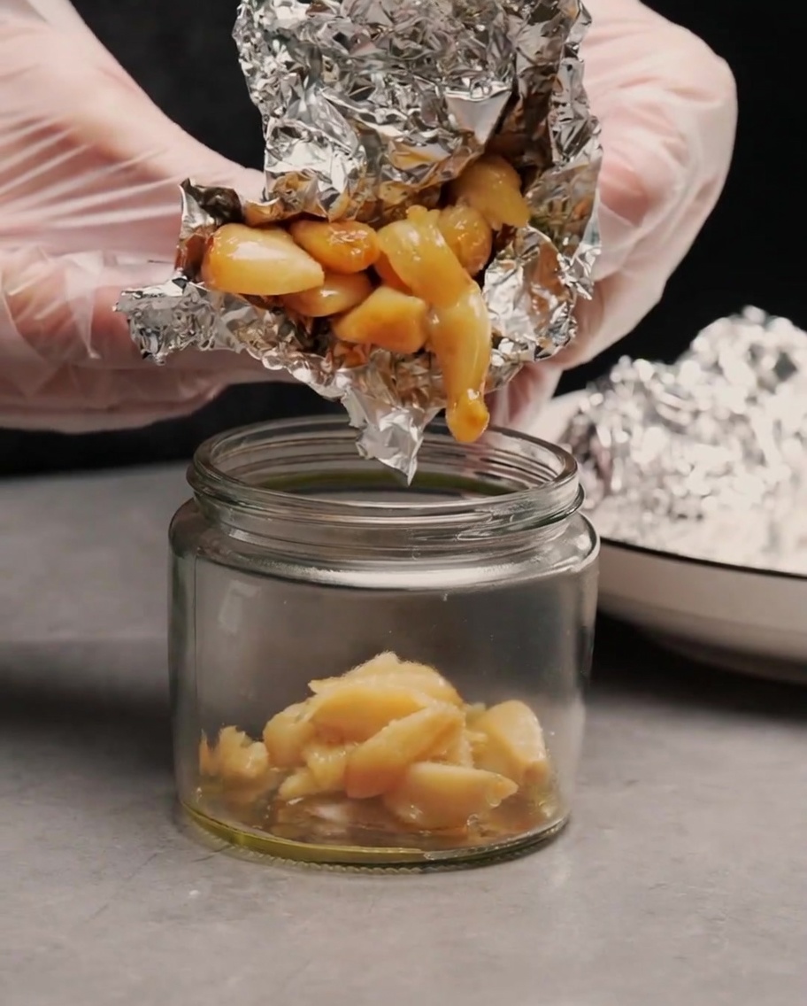 Manos con guantes exprimiendo dientes de ajo asados suaves y dorados fuera del papel de aluminio hacia un pequeño frasco de vidrio.