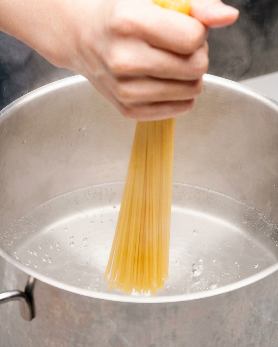 Una mano empujando un manojo de espaguetis secos directamente hacia una olla de acero inoxidable con agua hirviendo.
