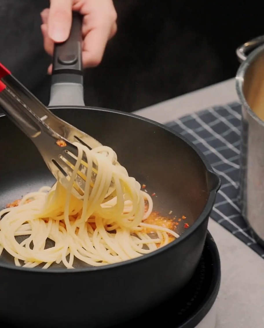 Espaguetis cocidos siendo mezclados con pinzas en una sartén con aceite de ajo y chile.