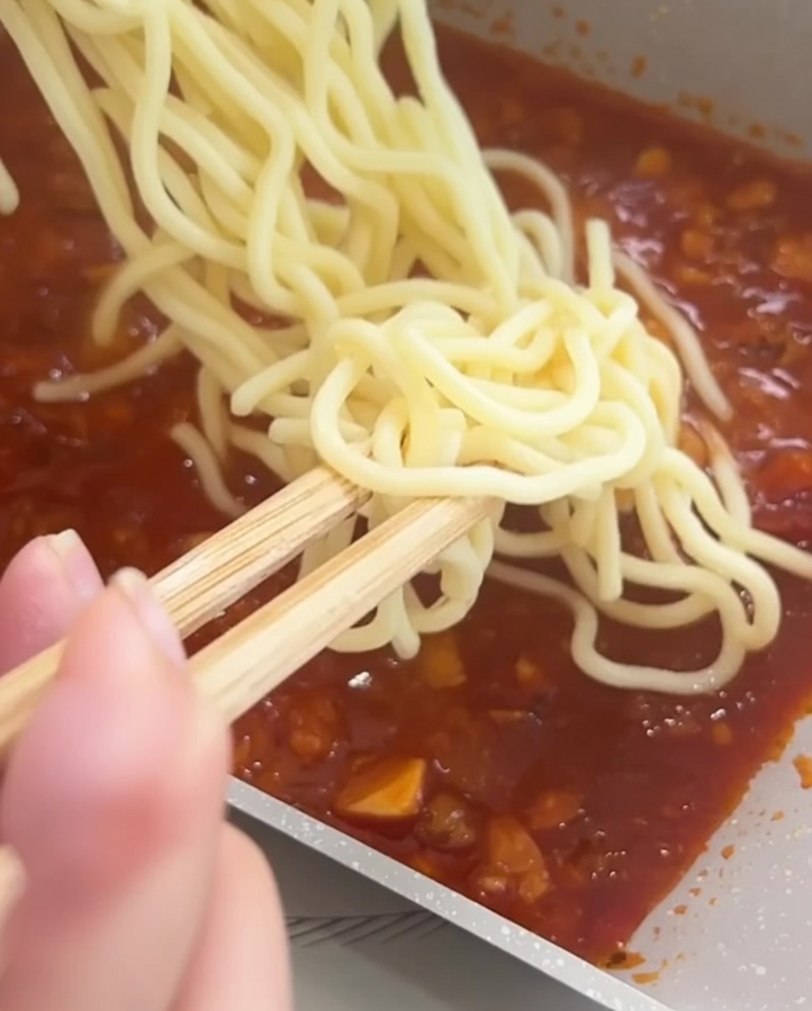 Espaguetis mezclándose con la salsa de carne y tomate en una sartén usando unos palillos.