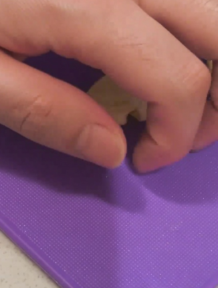 Primer plano de una mano presionando la parte plana de un cuchillo sobre un diente de ajo en una tabla de cortar morada.