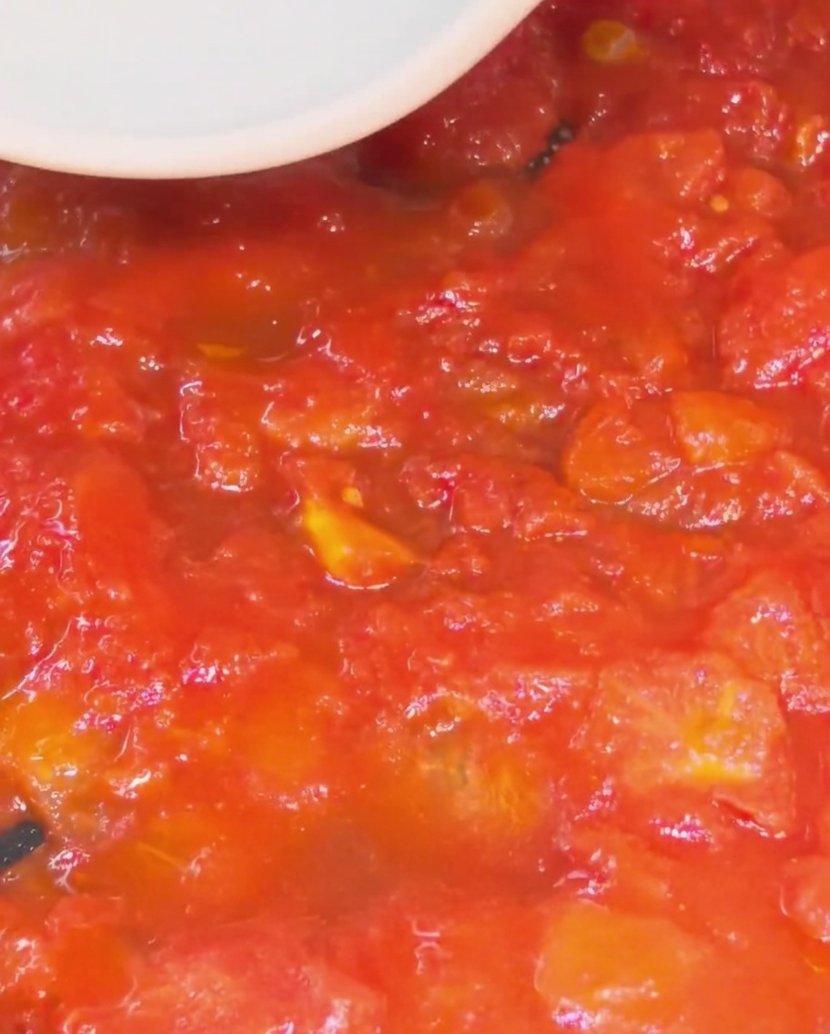 Primer plano de una espátula de silicona blanca presionando y triturando trozos de tomate rojo para formar una salsa espesa y suave en una sartén.