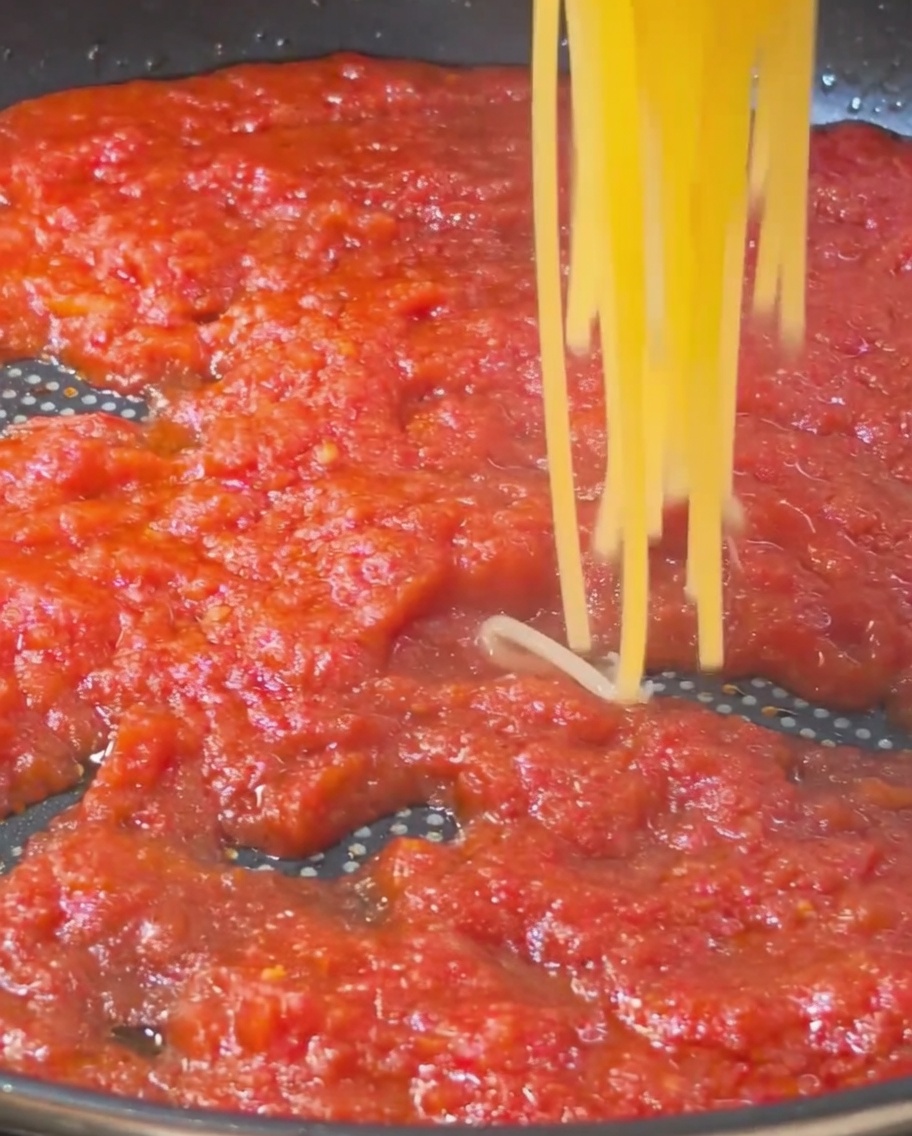 Una porción de espaguetis dorados cocidos siendo levantados y colocados en una sartén llena de una rica salsa de tomate roja.