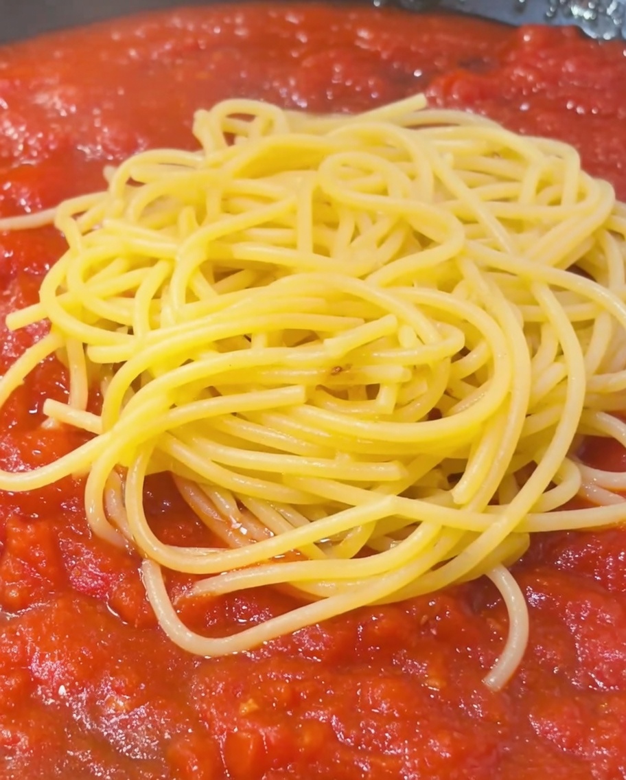 Una cucharada de agua con almidón de la pasta rociándose sobre una pila de espaguetis y salsa de tomate en una sartén.